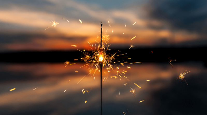 A lit sparkler glows, emitting sparks in front of a lake at sunset.